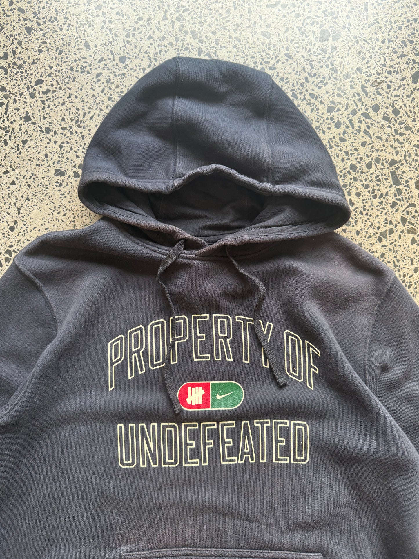 2010s Nike Undefeated hoodie size M laid on polished concrete background — black with bold graphics.