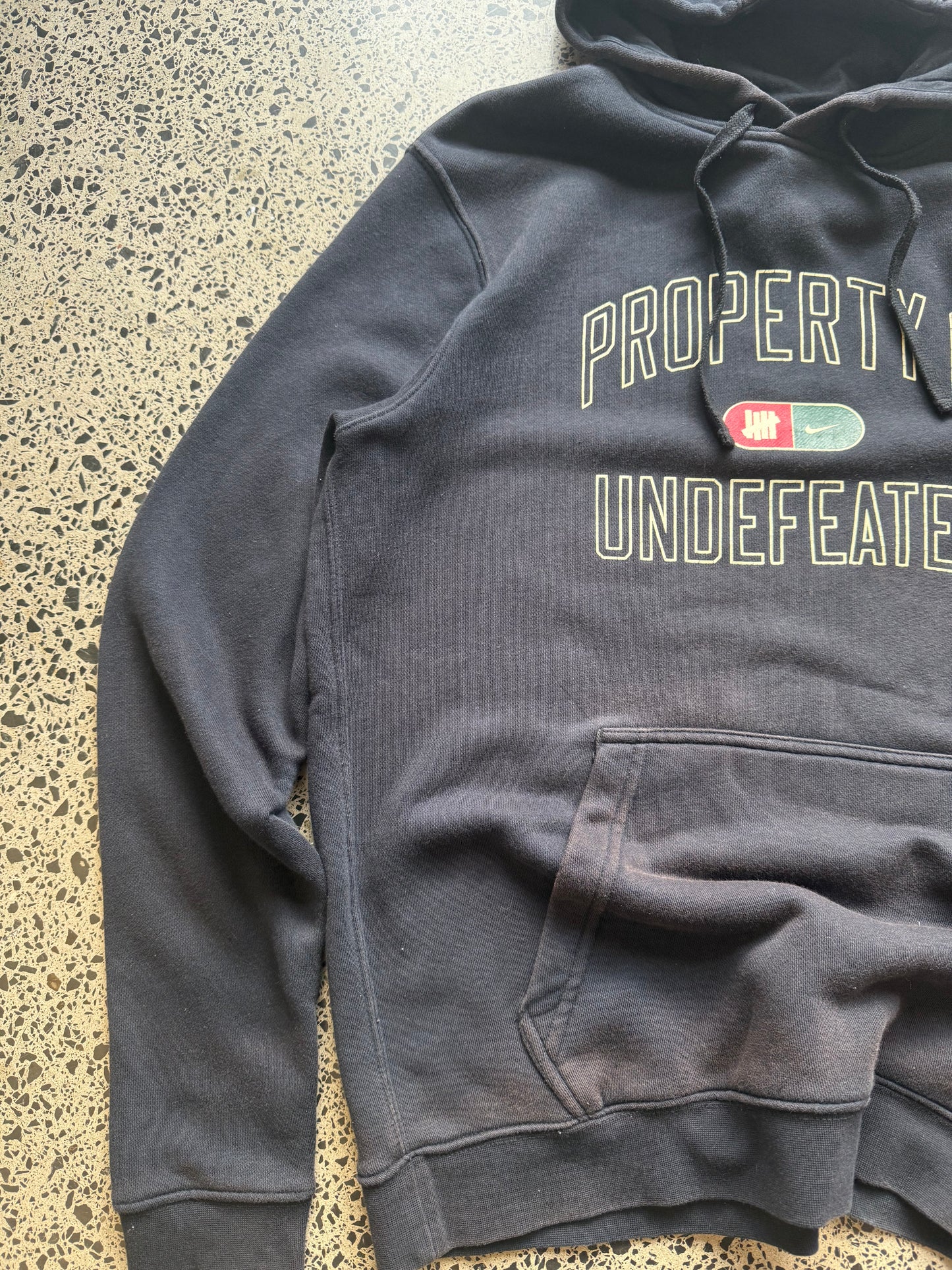 2010s Nike Undefeated hoodie size M laid on polished concrete background — black with bold graphics.
