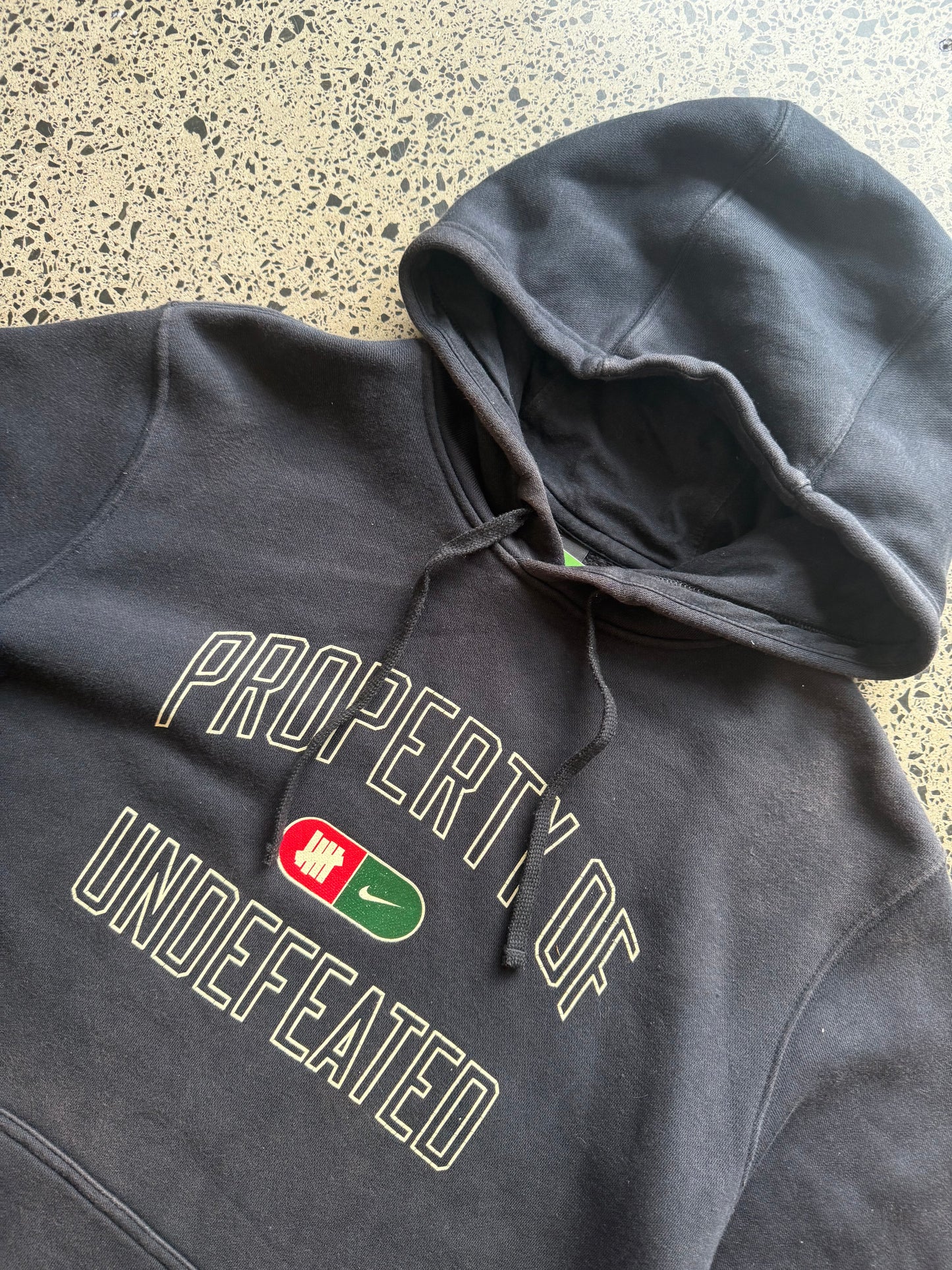 2010s Nike Undefeated hoodie size M laid on polished concrete background — black with bold graphics.