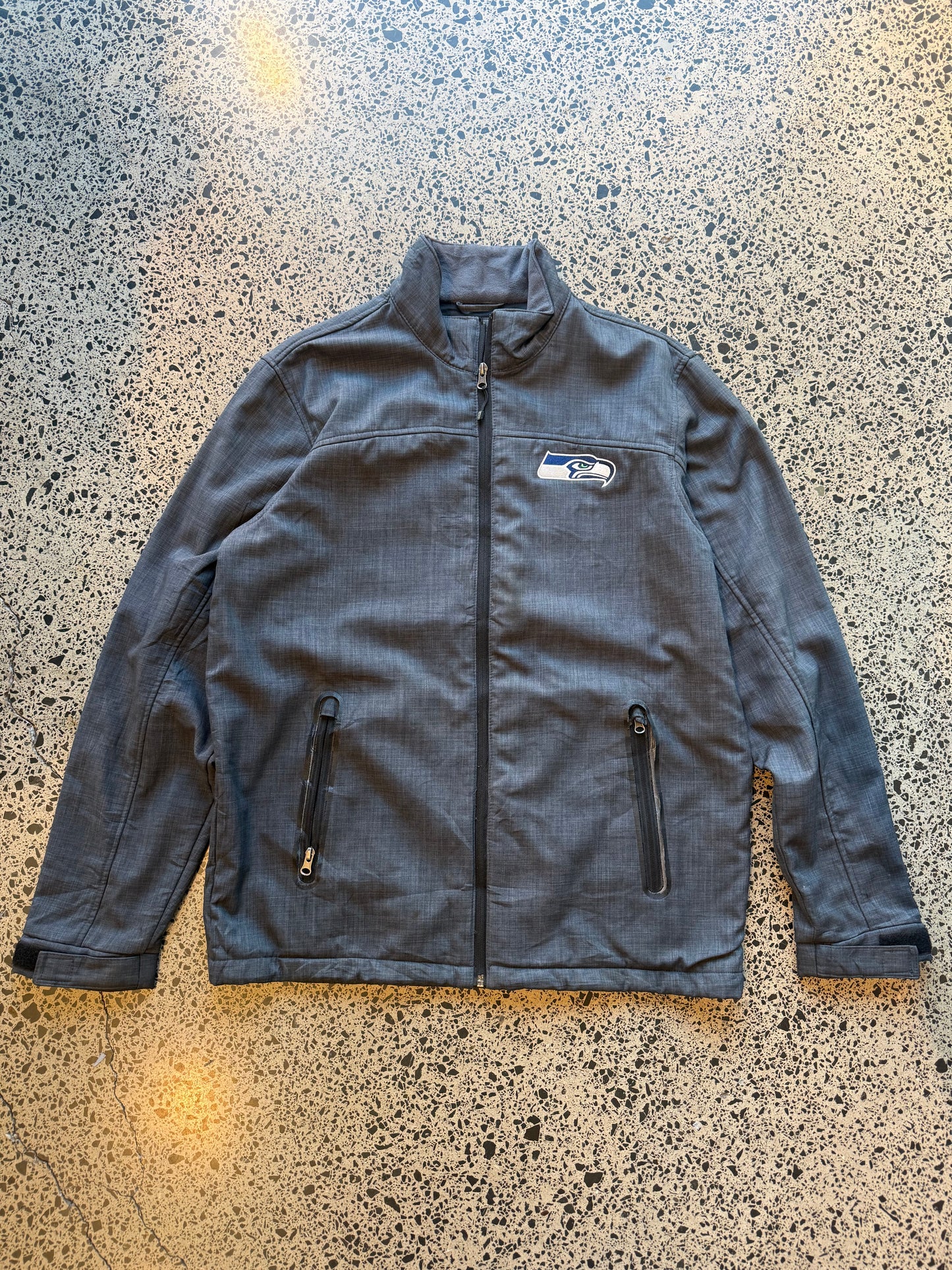 Seattle Seahawks NFL soft shell jacket size M laid flat on polished concrete background, lightweight sportswear jacket with subtle team branding and clean fit.
