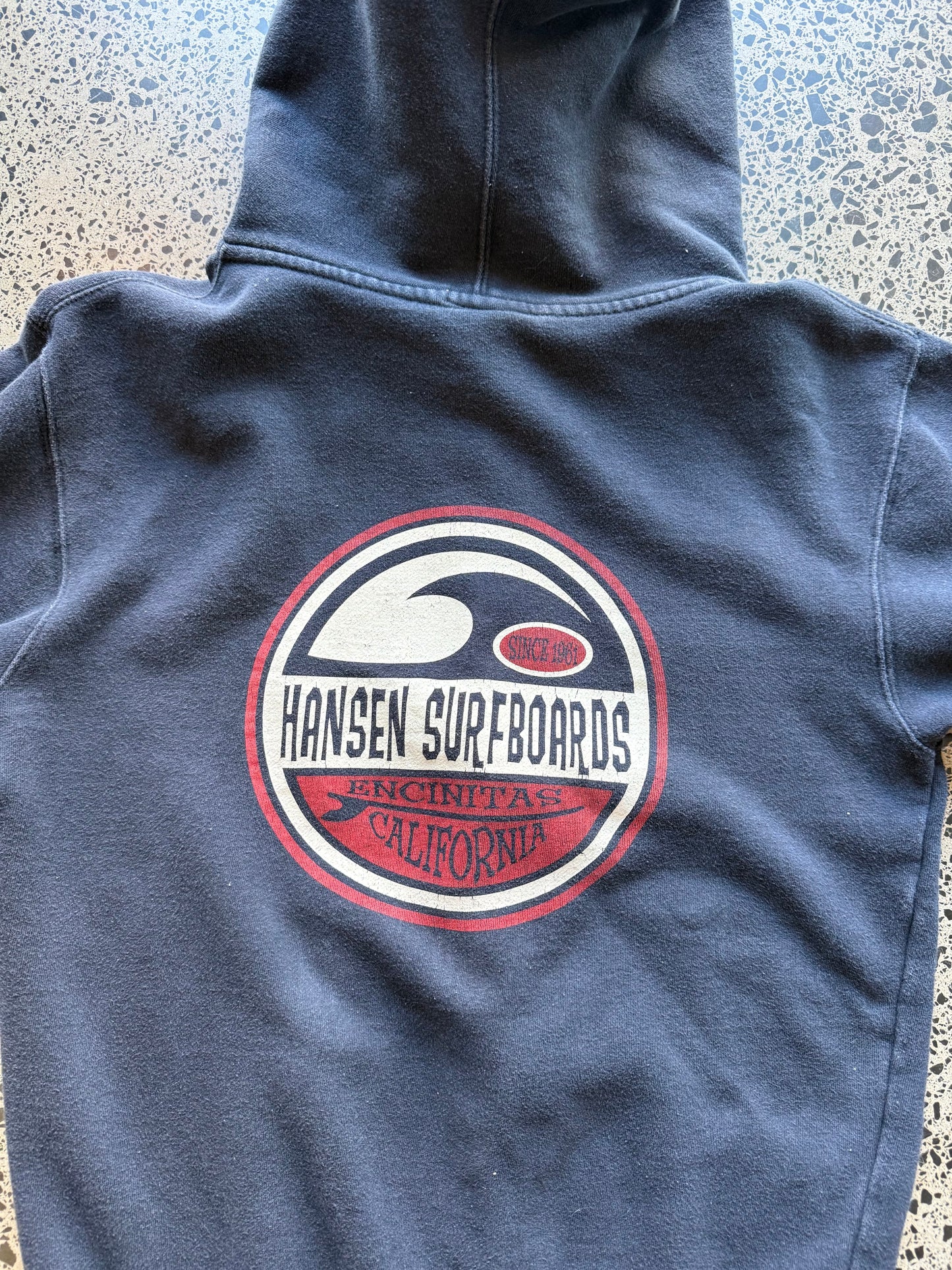 Hansen Surfboards California zip-up hoodie size M laid flat on polished concrete background, surfwear hoodie with minimal branding and relaxed fit.