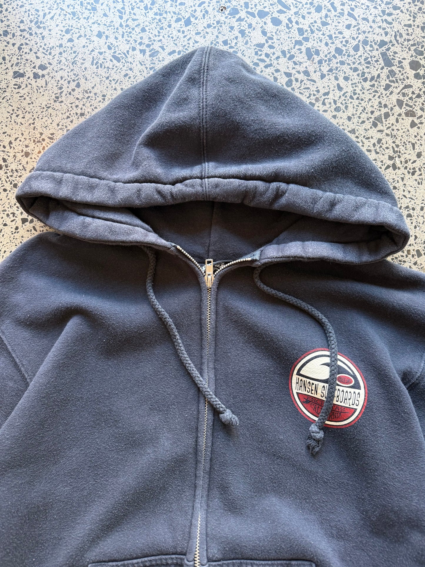 Hansen Surfboards California zip-up hoodie size M laid flat on polished concrete background, surfwear hoodie with minimal branding and relaxed fit.