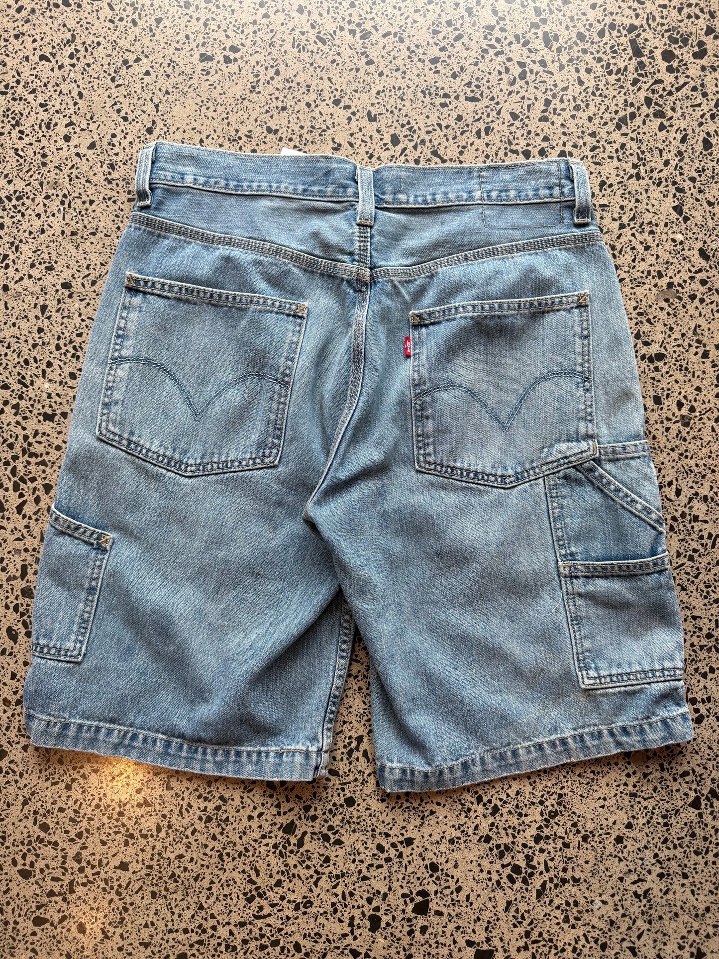 Levi's light blue wash denim jorts, size 32W, laid on polished concrete background. Knee cut with classic stitching and soft wash