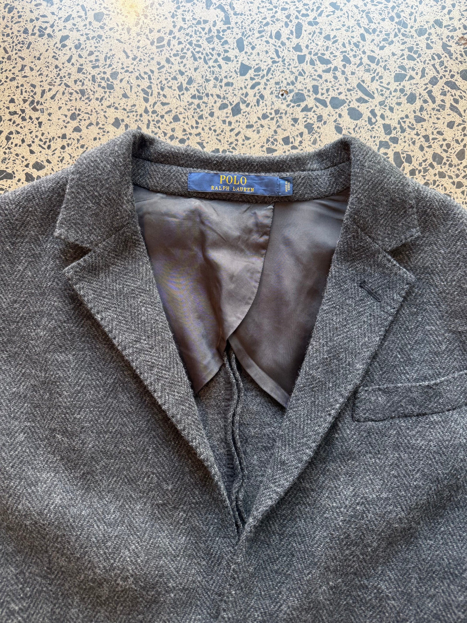 Ralph Lauren herringbone 100 percent wool blazer jacket laid flat on polished concrete background, classic tailored jacket with textured pattern and clean fit.