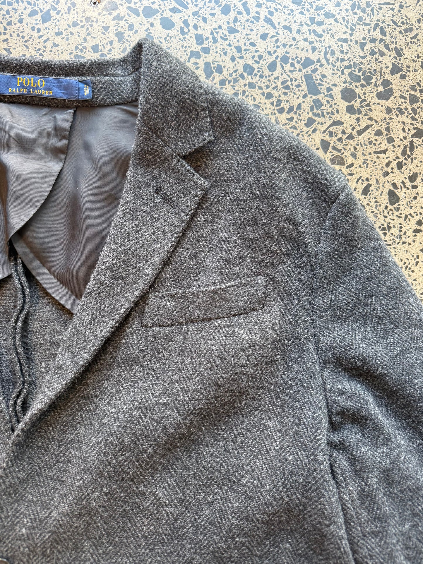 Ralph Lauren herringbone 100 percent wool blazer jacket laid flat on polished concrete background, classic tailored jacket with textured pattern and clean fit.