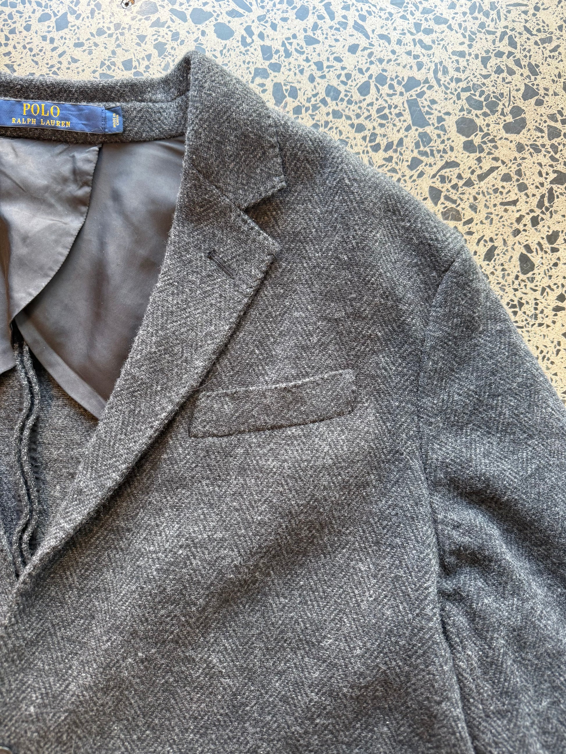 Ralph Lauren herringbone 100 percent wool blazer jacket laid flat on polished concrete background, classic tailored jacket with textured pattern and clean fit.