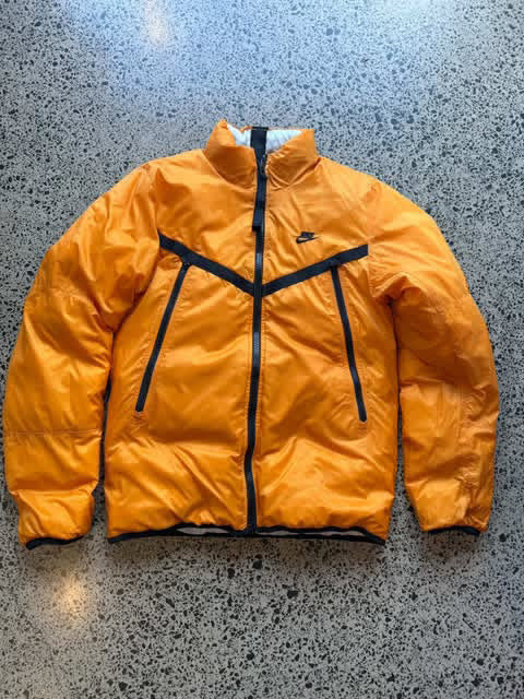 Reversible orange and white Nike jacket laid flat on a polished concrete background featuring classic Nike branding and dual-colour design