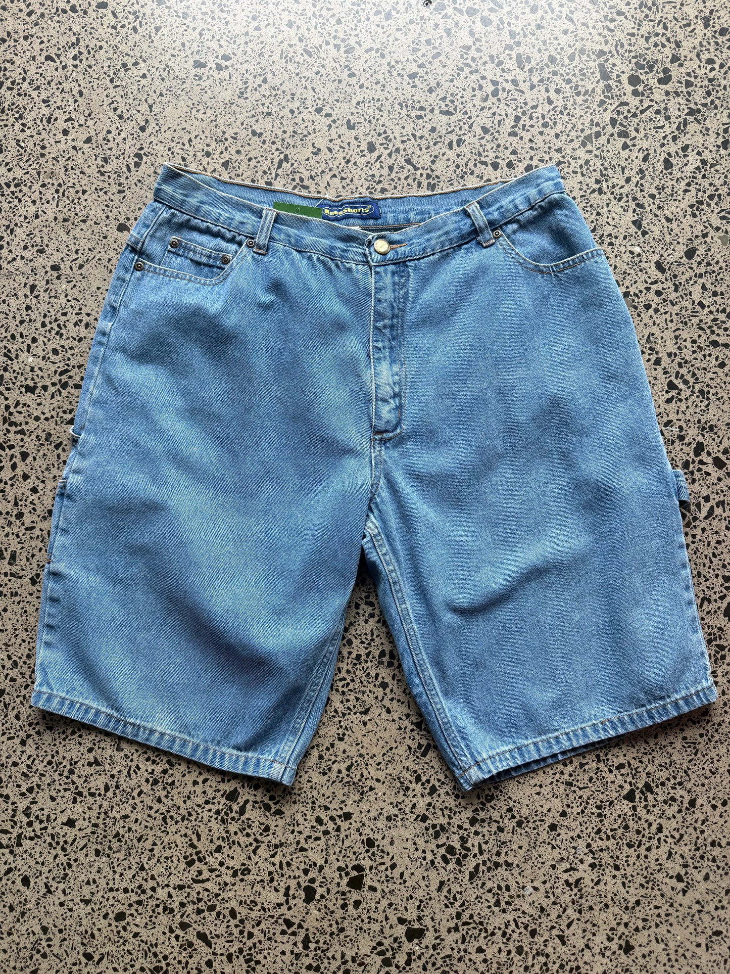 Kneeshorts  light-wash denim jorts, size 30W, laid on polished concrete background. Just above knee cut with classic stitching and soft wash