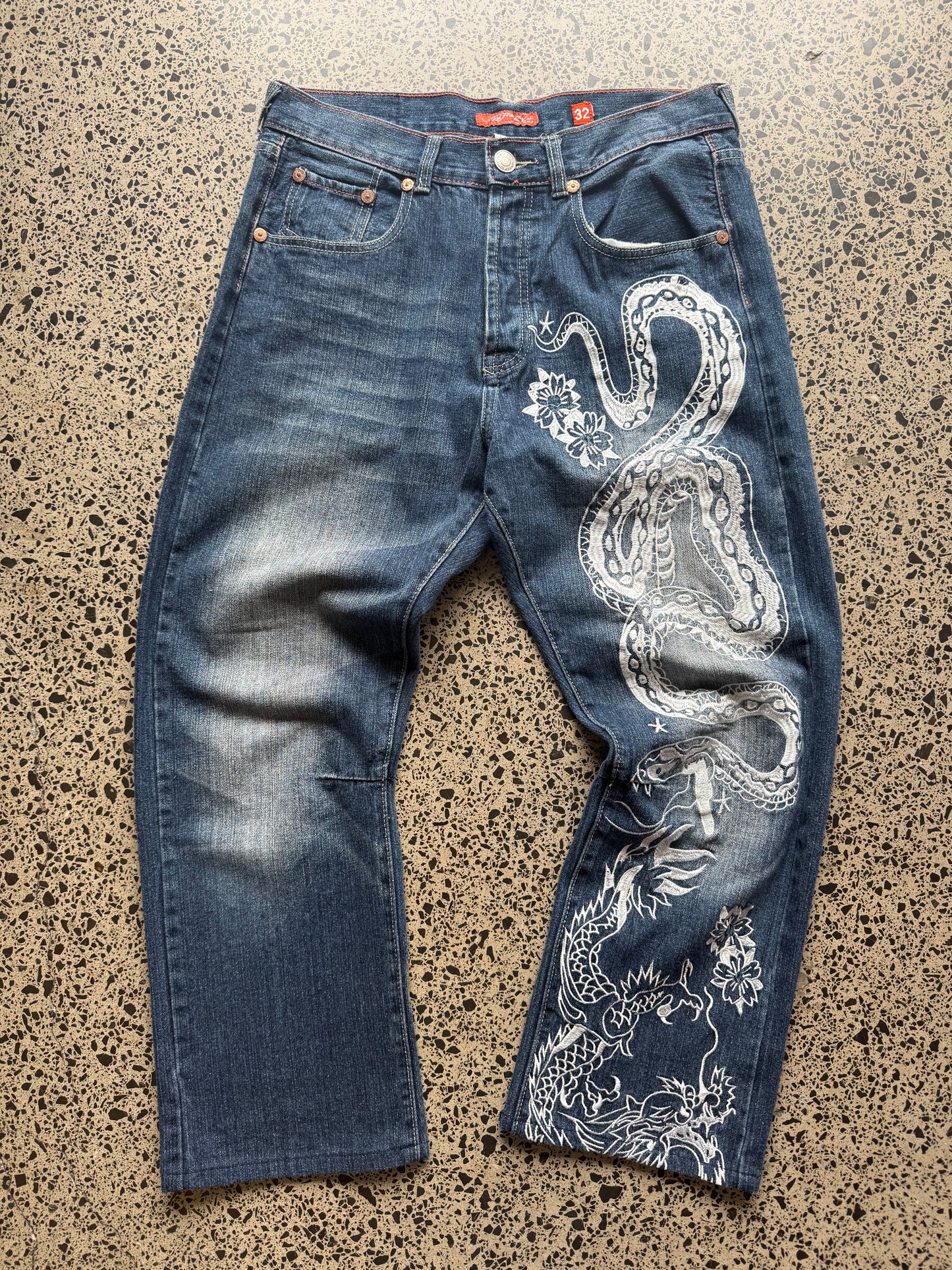 2008 Ed Hardy Snake and Dragon embroidered denim jeans with rhinestone detailing laid flat on a polished concrete background.