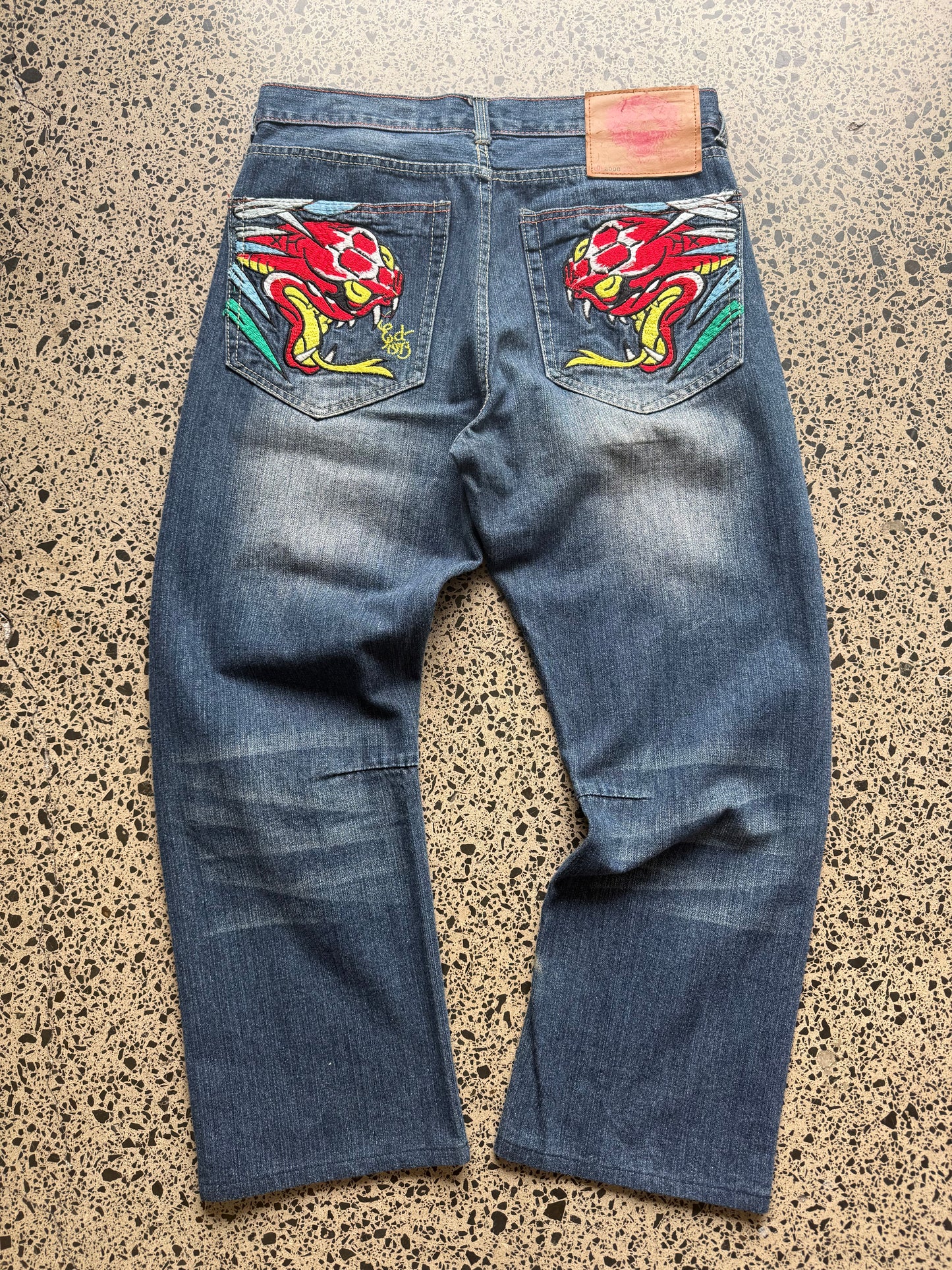 2008 Ed Hardy Snake and Dragon embroidered denim jeans with rhinestone detailing laid flat on a polished concrete background.