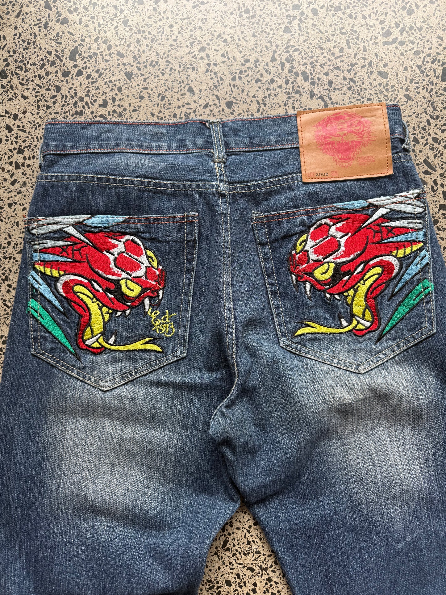 2008 Ed Hardy Snake and Dragon embroidered denim jeans with rhinestone detailing laid flat on a polished concrete background.