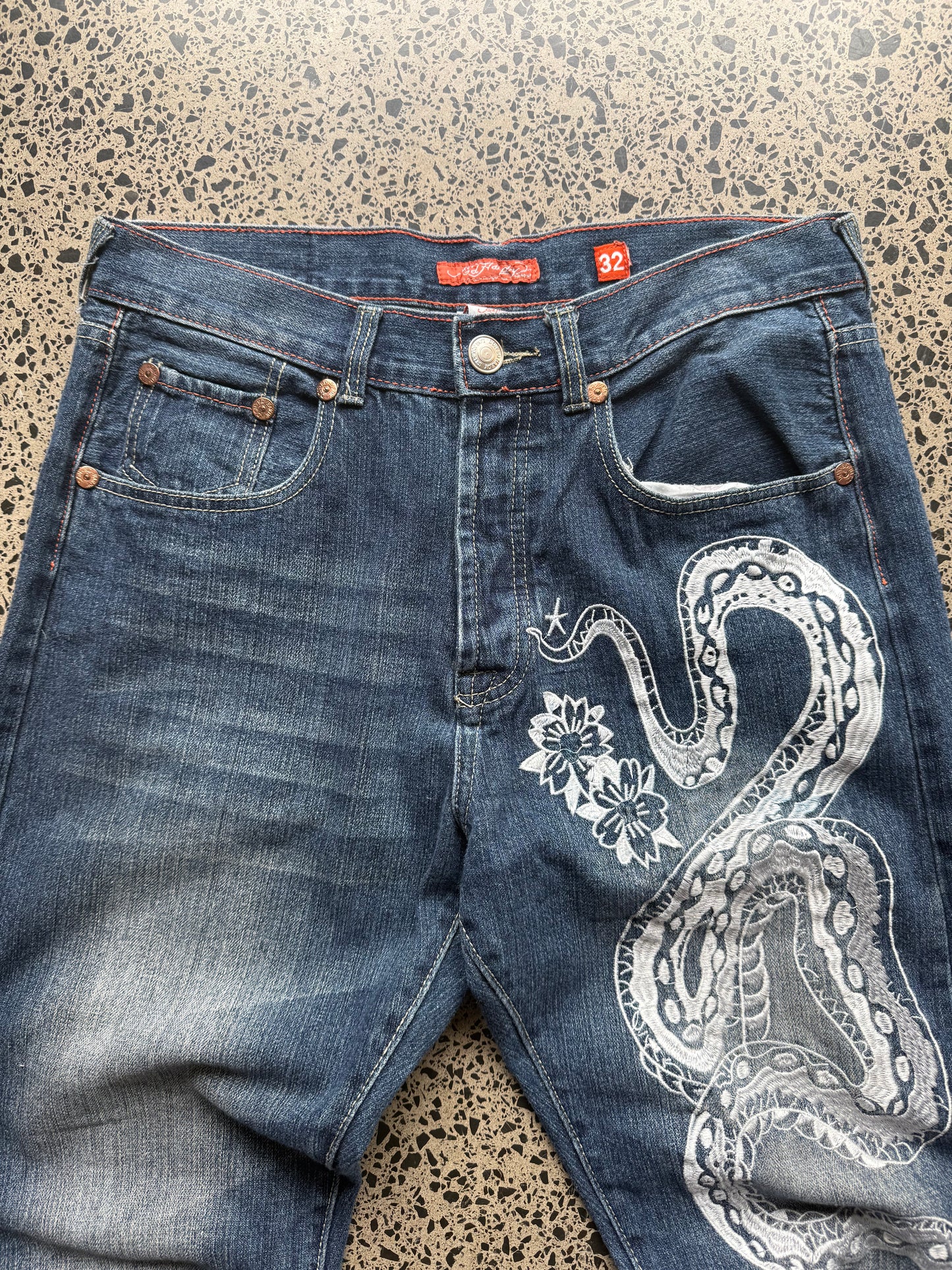 2008 Ed Hardy Snake and Dragon embroidered denim jeans with rhinestone detailing laid flat on a polished concrete background.