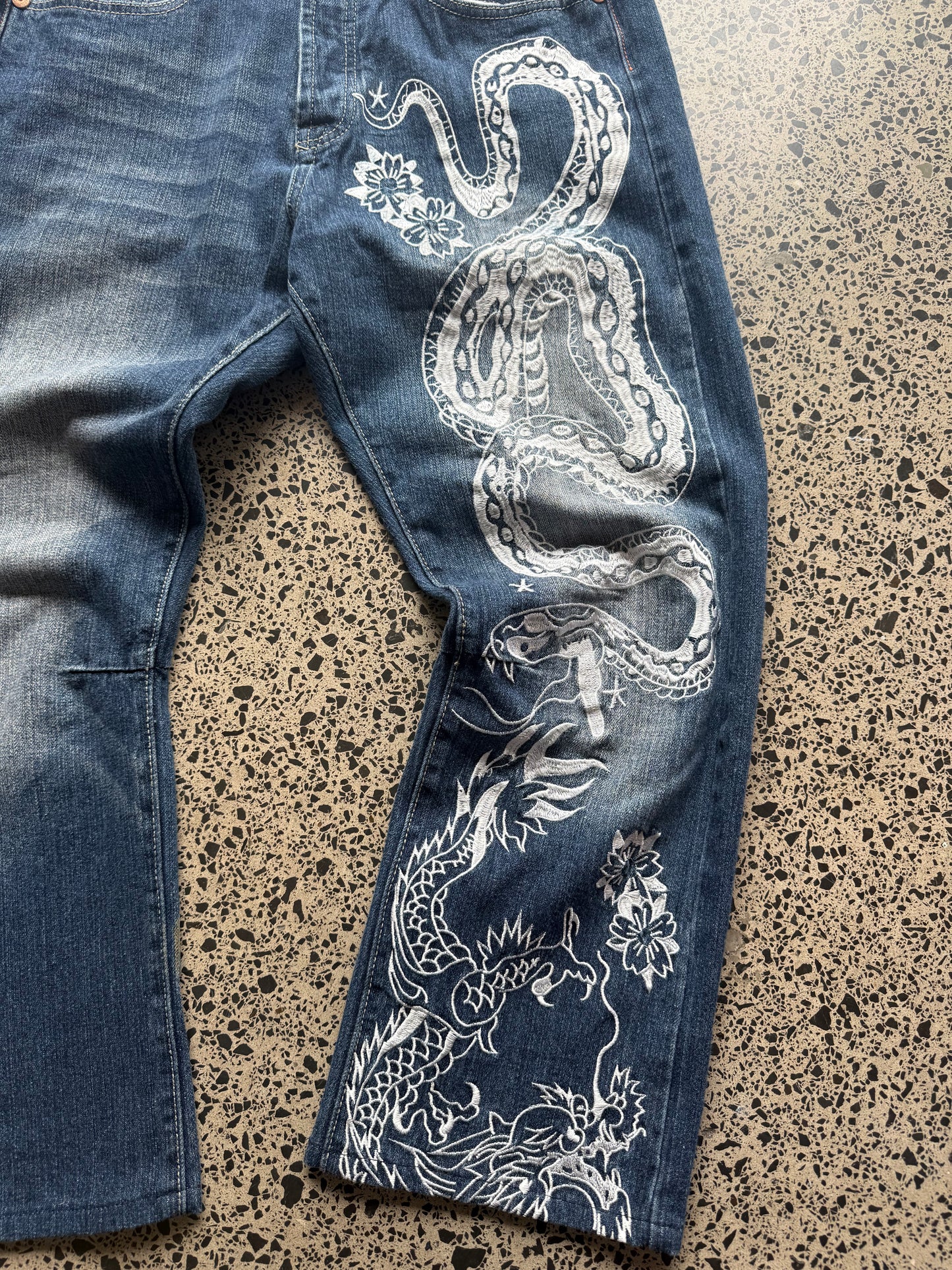 2008 Ed Hardy Snake and Dragon embroidered denim jeans with rhinestone detailing laid flat on a polished concrete background.
