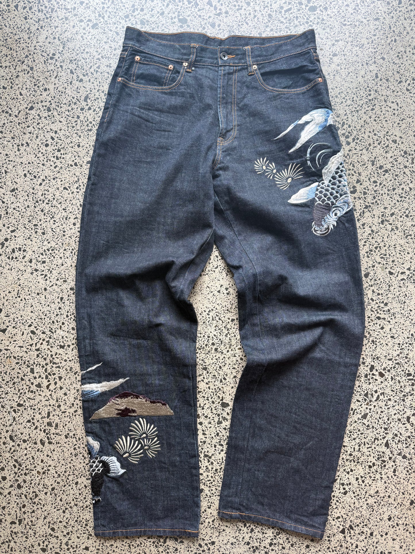 Karakuri Tamashii embroidered denim jeans with dragon and wave detailing laid flat on a polished concrete background.