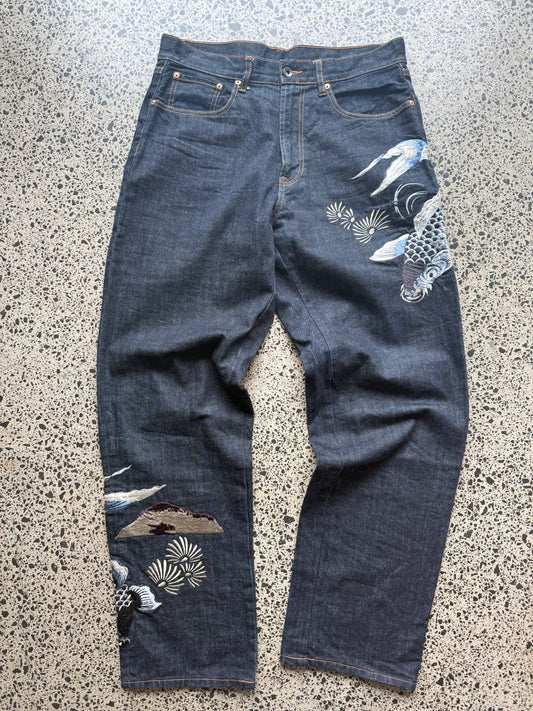 Karakuri Tamashii embroidered denim jeans with dragon and wave detailing laid flat on a polished concrete background.