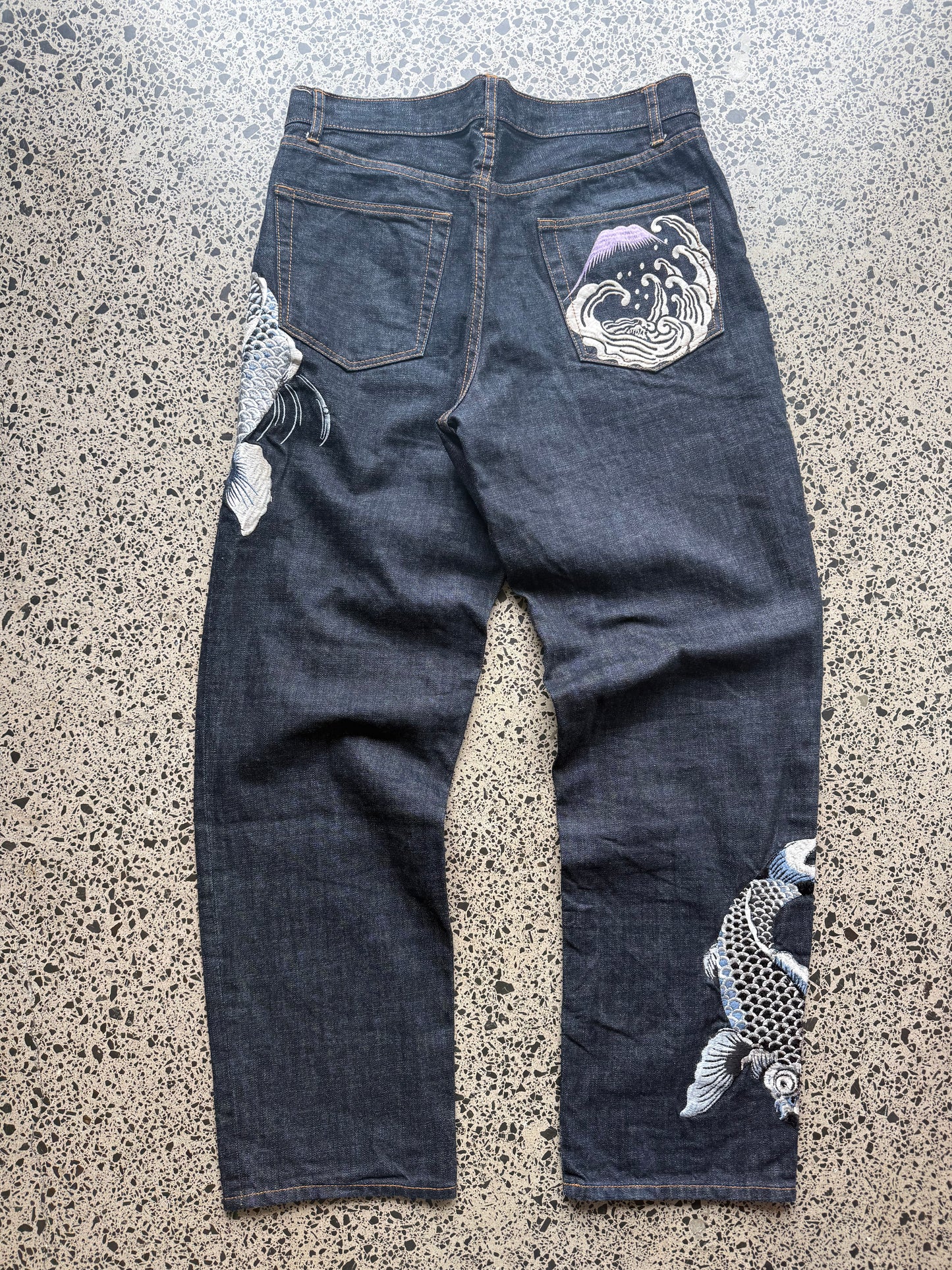 Karakuri Tamashii embroidered denim jeans with dragon and wave detailing laid flat on a polished concrete background.
