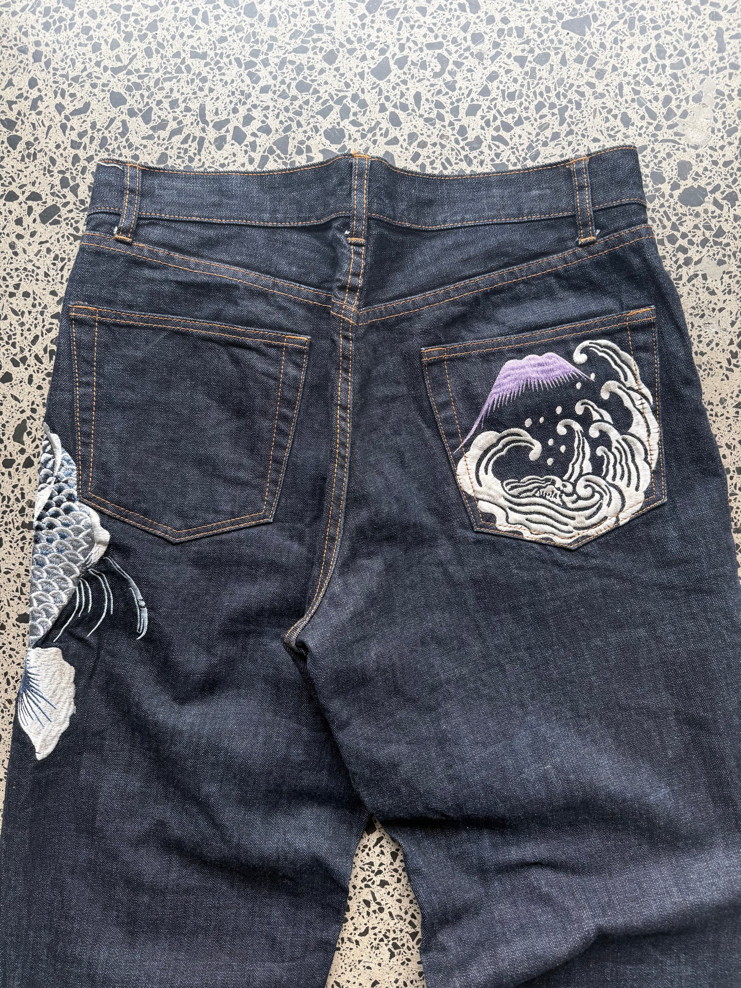 Karakuri Tamashii embroidered denim jeans with dragon and wave detailing laid flat on a polished concrete background.