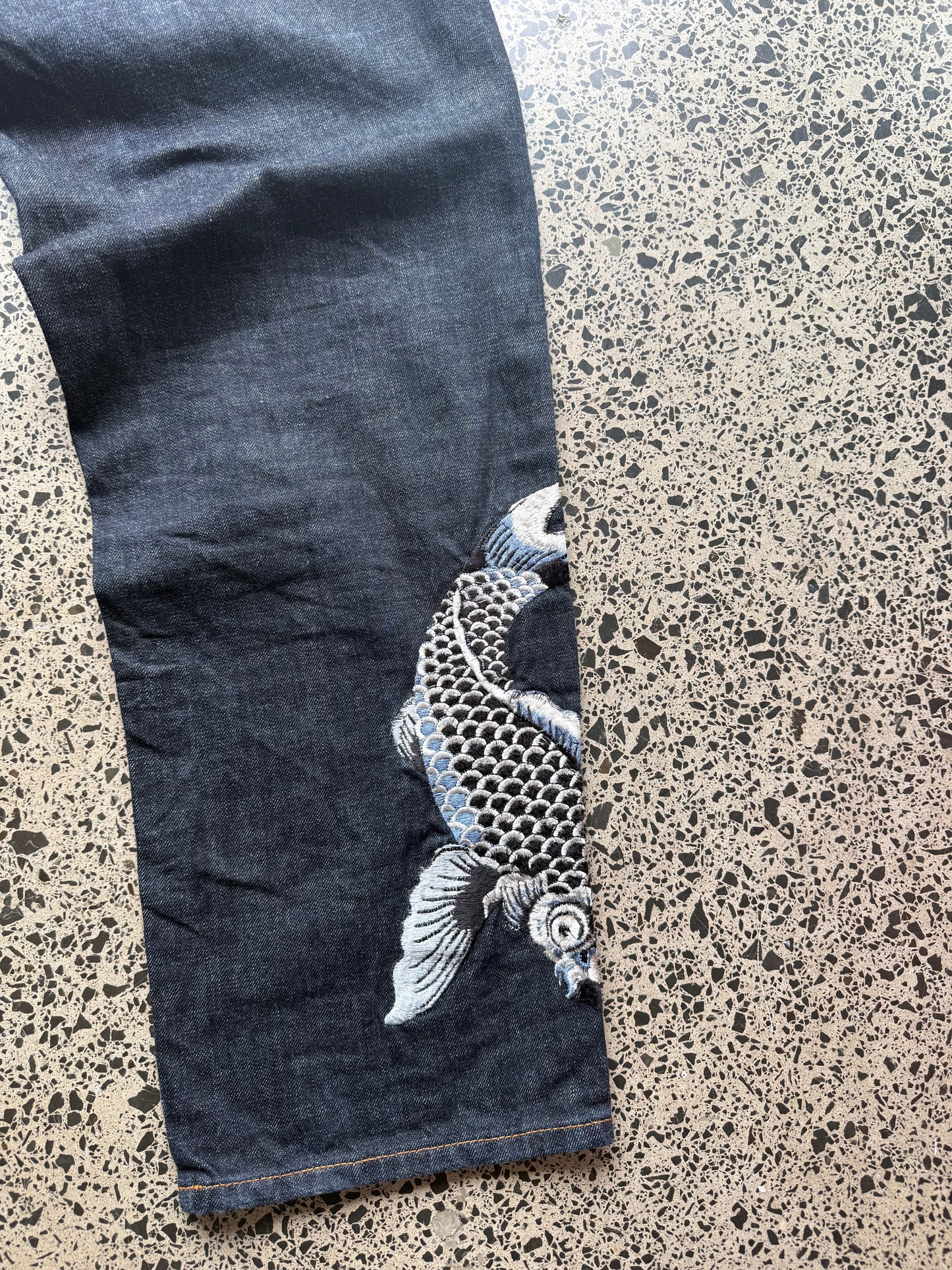 Karakuri Tamashii embroidered denim jeans with dragon and wave detailing laid flat on a polished concrete background.