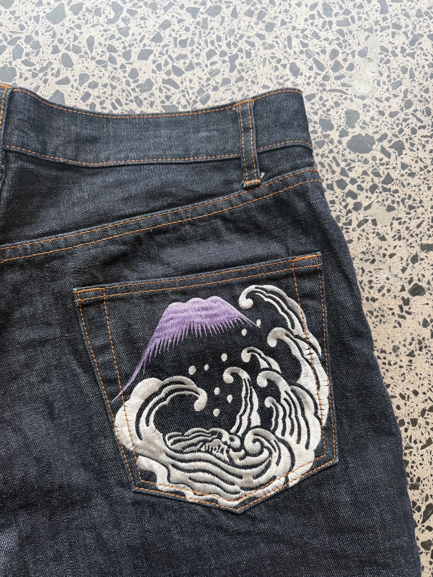Karakuri Tamashii embroidered denim jeans with dragon and wave detailing laid flat on a polished concrete background.