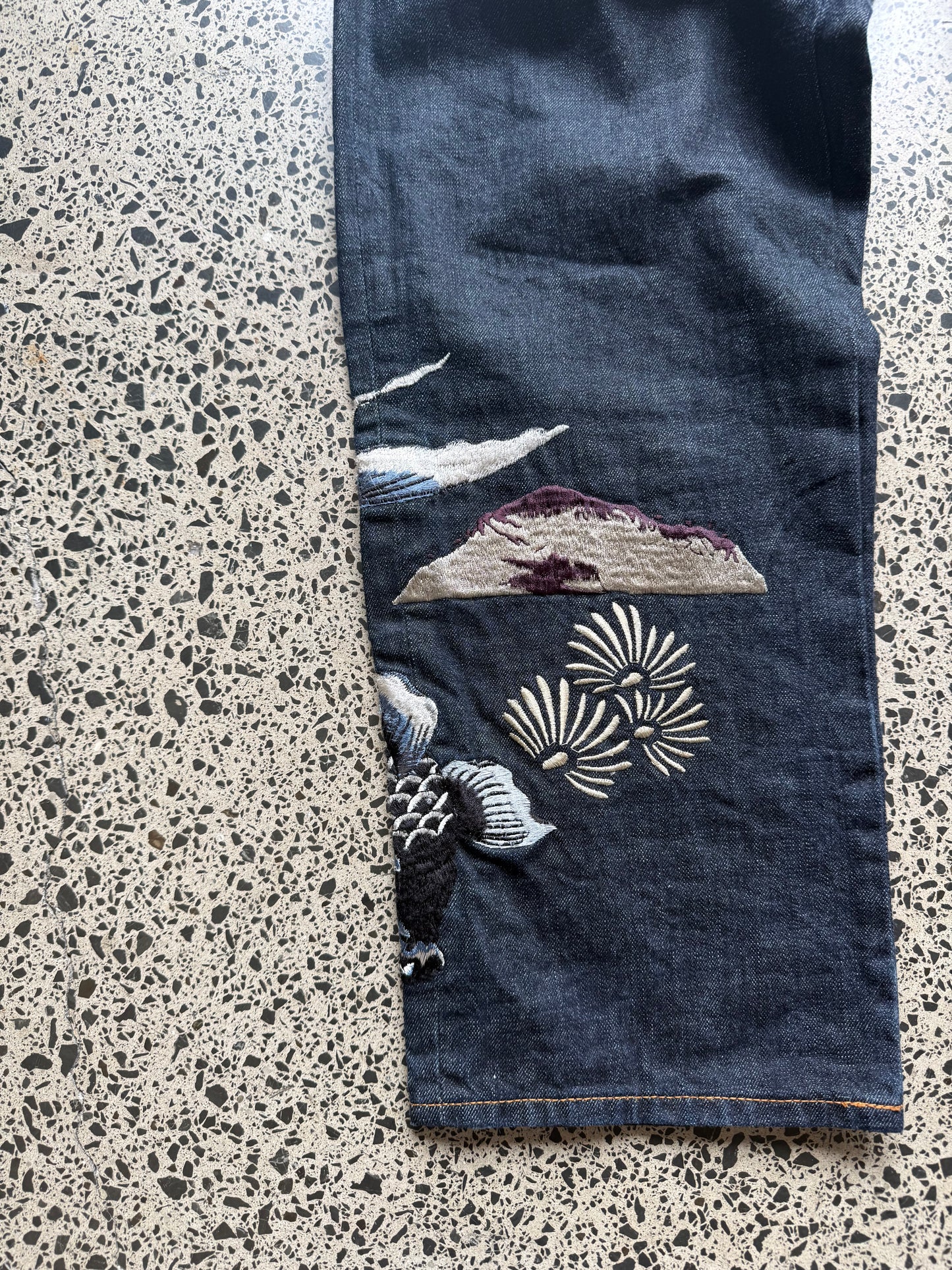 Karakuri Tamashii embroidered denim jeans with dragon and wave detailing laid flat on a polished concrete background.