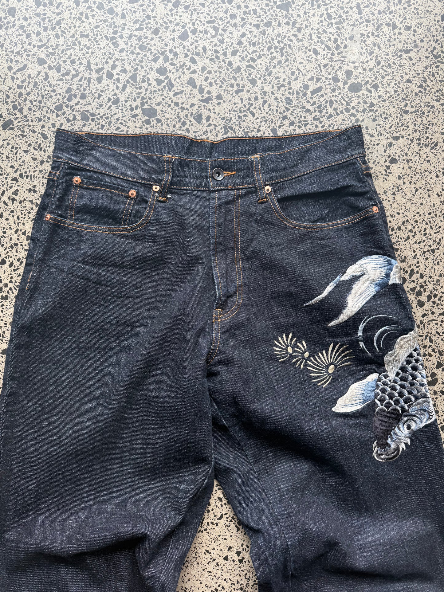 Karakuri Tamashii embroidered denim jeans with dragon and wave detailing laid flat on a polished concrete background.