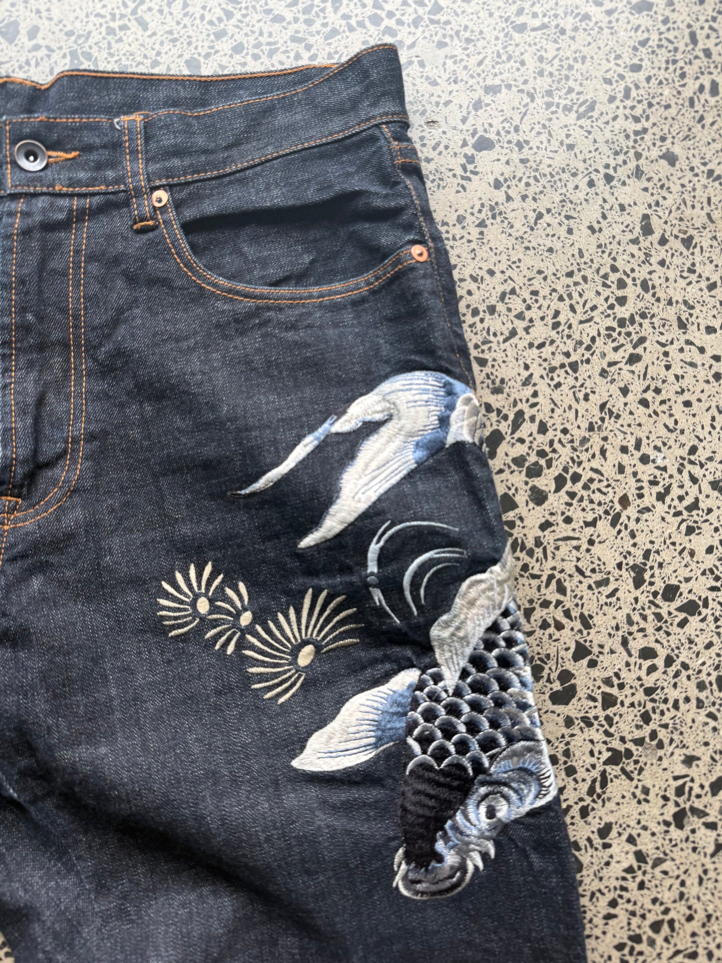 Karakuri Tamashii embroidered denim jeans with dragon and wave detailing laid flat on a polished concrete background.