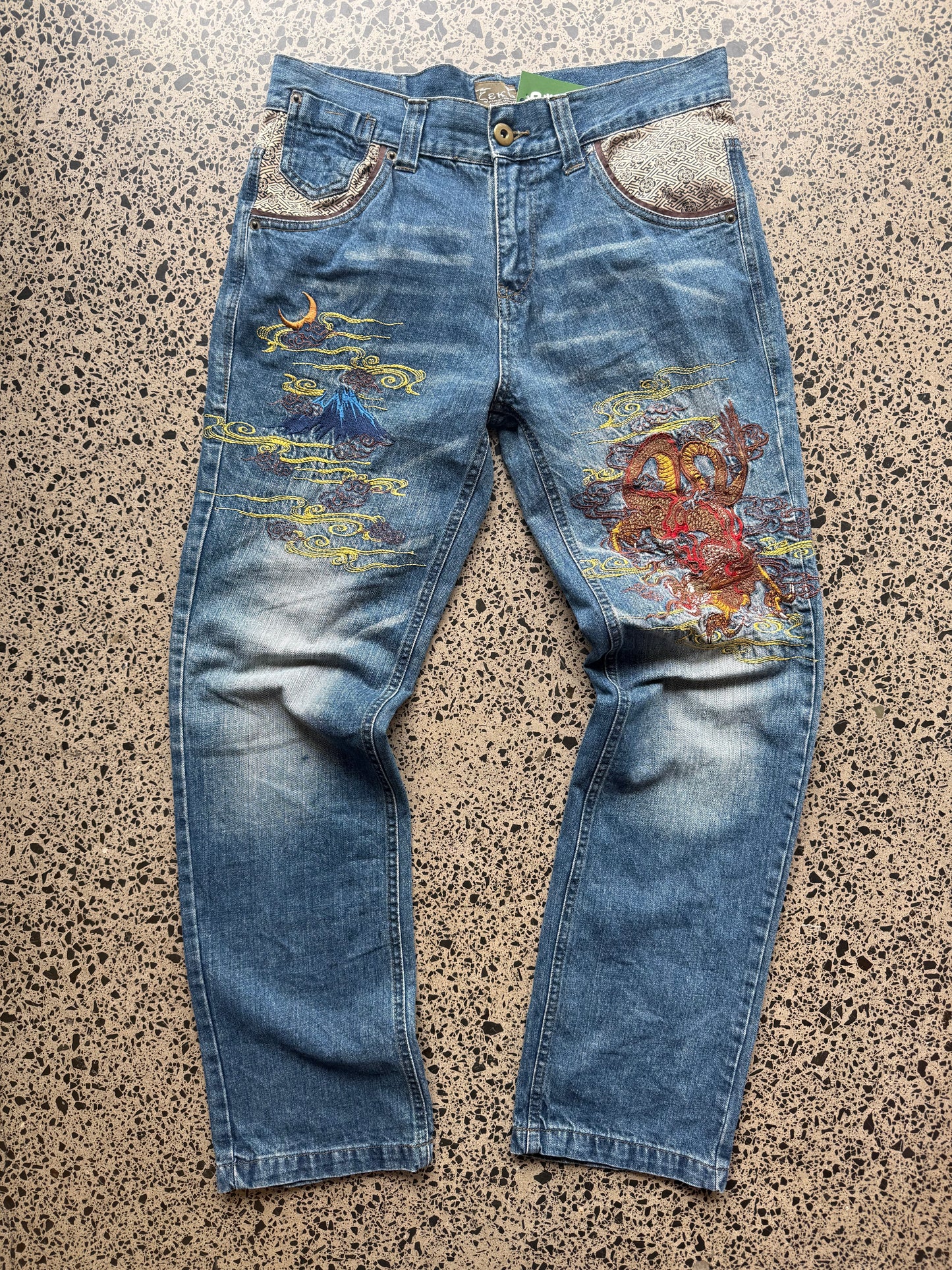 Seku light wash embroidered denim jeans with red and gold dragon detailing laid flat on a polished concrete background.