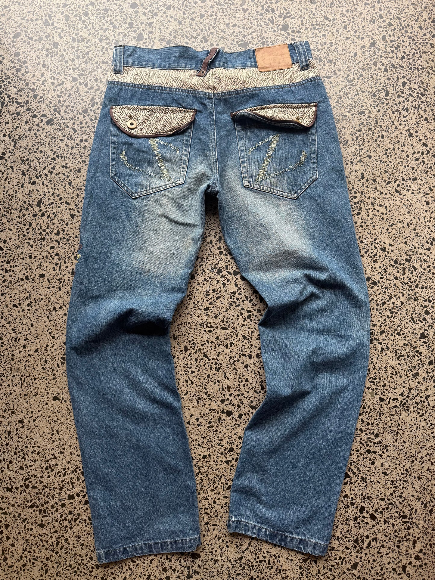 Seku light wash embroidered denim jeans with red and gold dragon detailing laid flat on a polished concrete background.