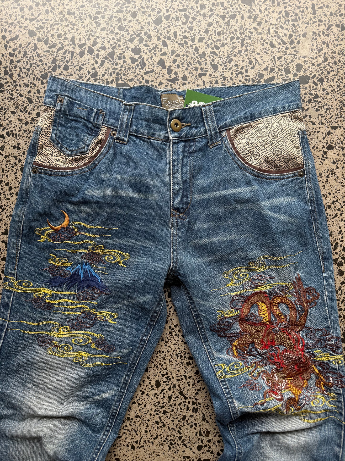 Seku light wash embroidered denim jeans with red and gold dragon detailing laid flat on a polished concrete background.