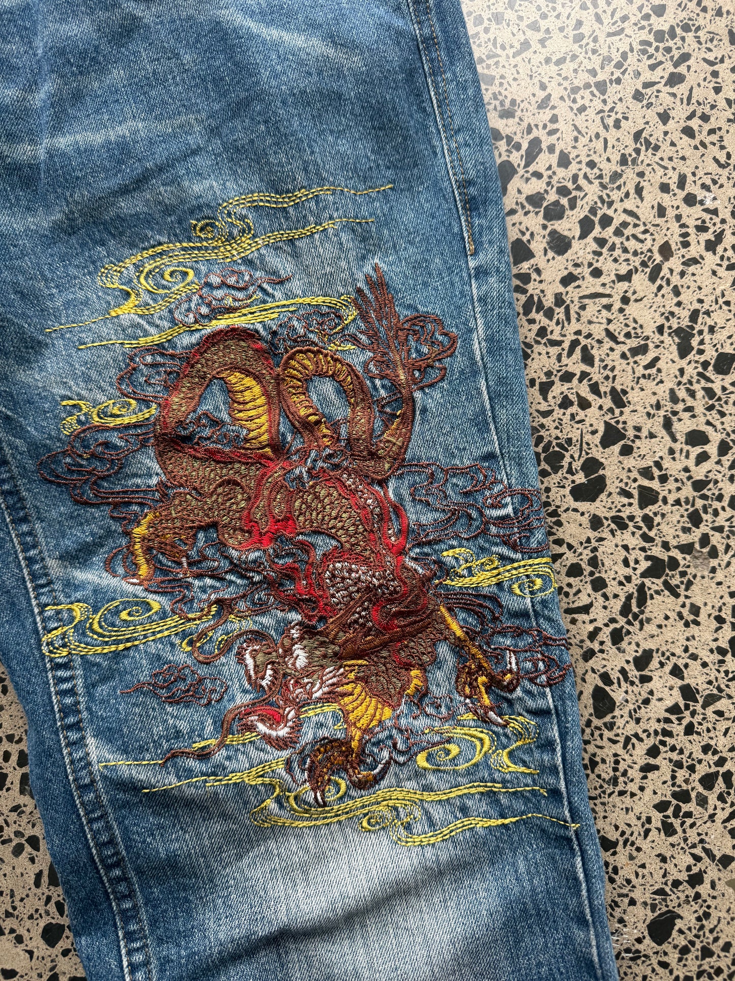 Seku light wash embroidered denim jeans with red and gold dragon detailing laid flat on a polished concrete background.