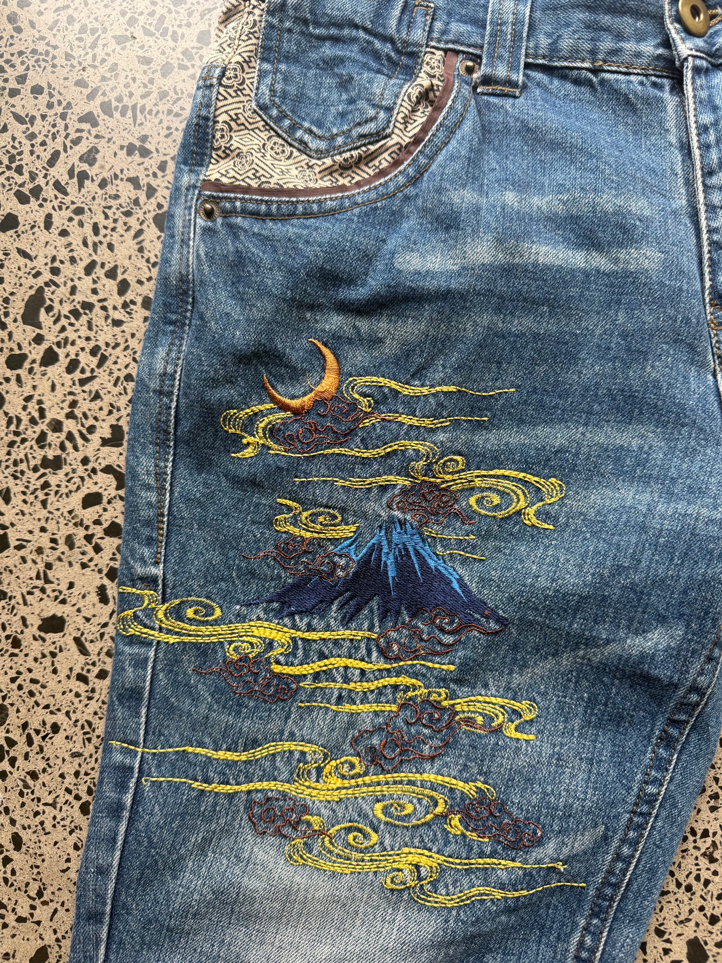 Seku light wash embroidered denim jeans with red and gold dragon detailing laid flat on a polished concrete background.