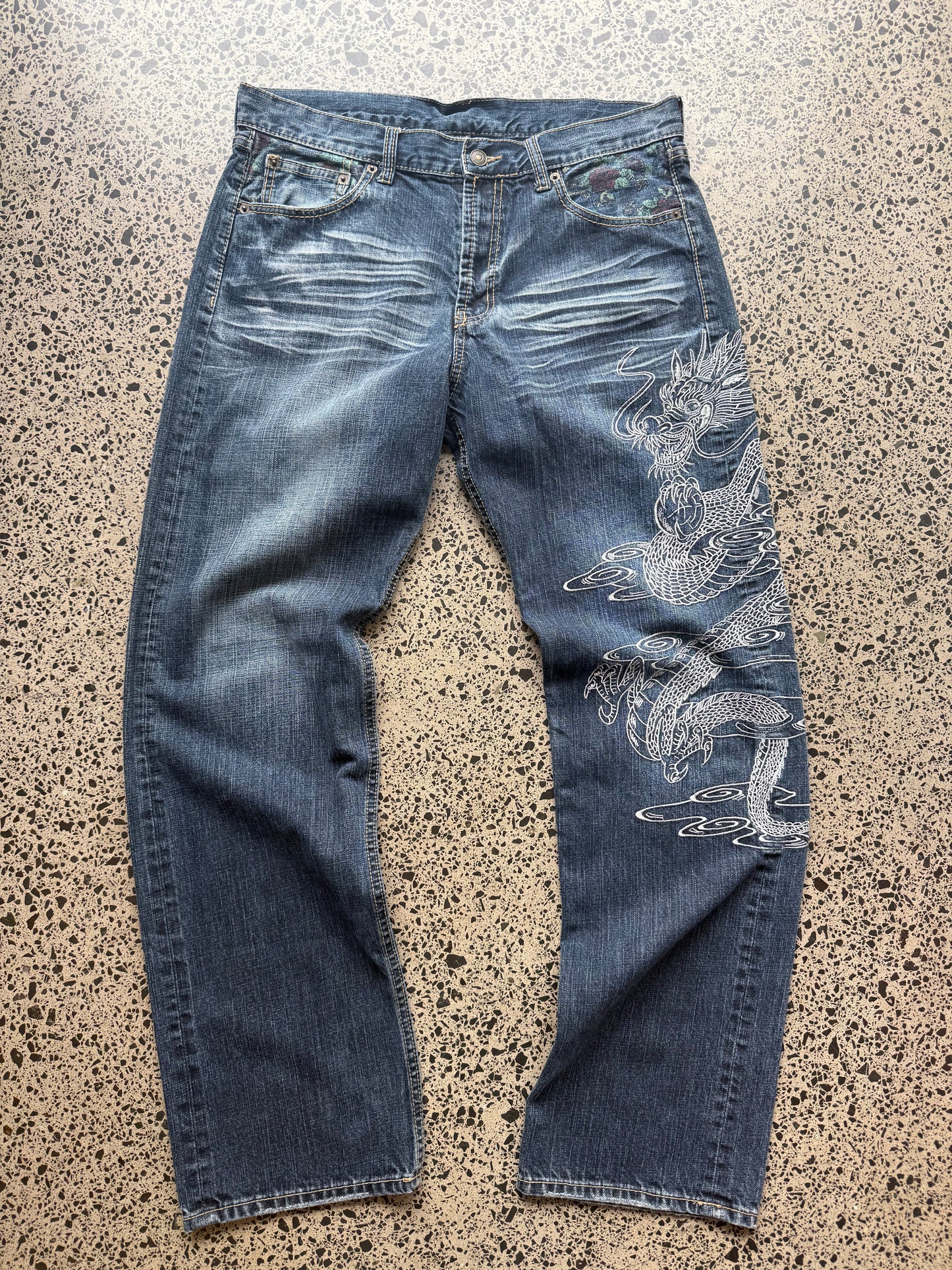 Un Deux dragon embroidered denim jeans, 34W x 34L, laid on polished concrete background — mid-blue wash with bold full-leg dragon embroidery.