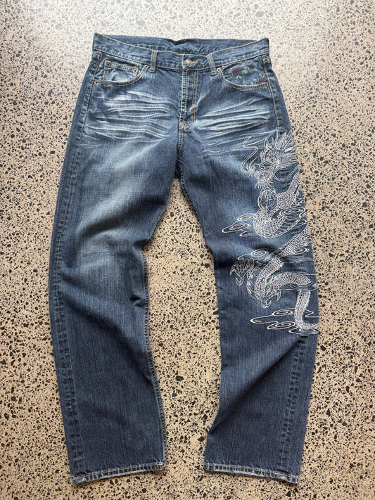Un Deux dragon embroidered denim jeans, 34W x 34L, laid on polished concrete background — mid-blue wash with bold full-leg dragon embroidery.