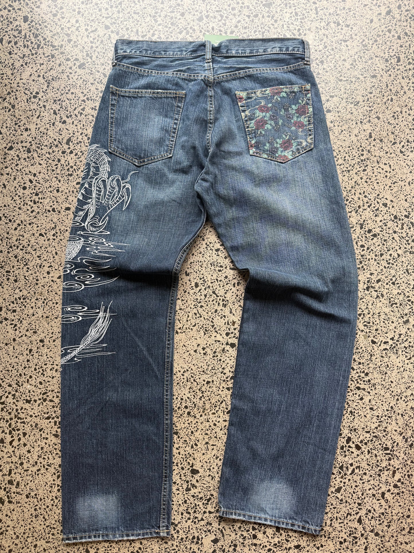 Un Deux dragon embroidered denim jeans, 34W x 34L, laid on polished concrete background — mid-blue wash with bold full-leg dragon embroidery.