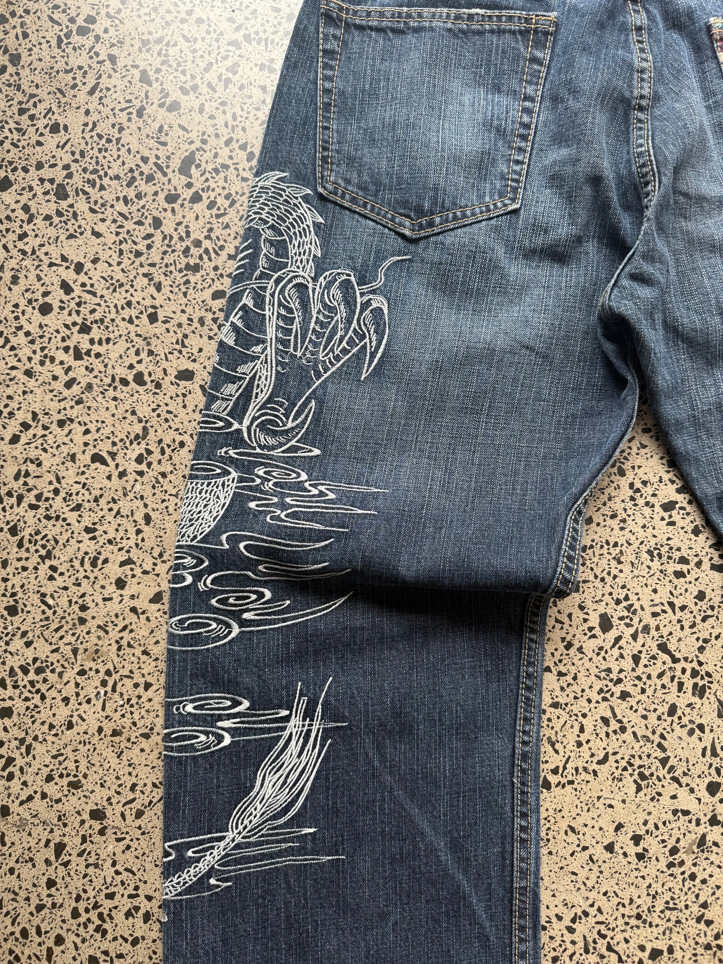 Un Deux dragon embroidered denim jeans, 34W x 34L, laid on polished concrete background — mid-blue wash with bold full-leg dragon embroidery.