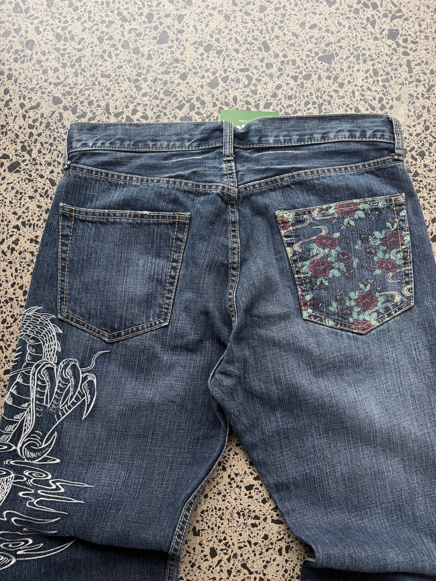 Un Deux dragon embroidered denim jeans, 34W x 34L, laid on polished concrete background — mid-blue wash with bold full-leg dragon embroidery.