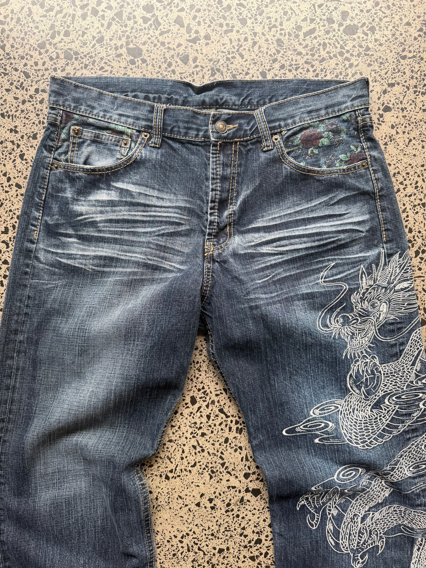 Un Deux dragon embroidered denim jeans, 34W x 34L, laid on polished concrete background — mid-blue wash with bold full-leg dragon embroidery.