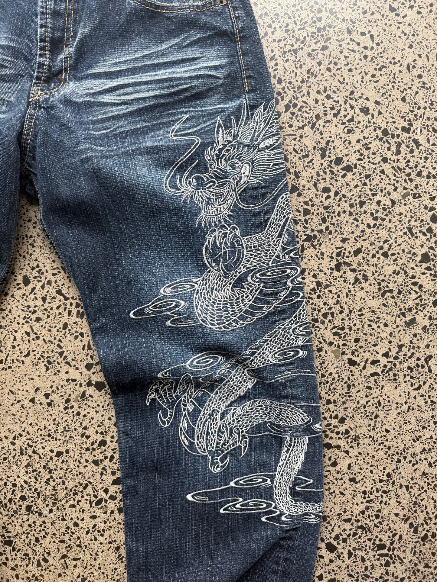 Un Deux dragon embroidered denim jeans, 34W x 34L, laid on polished concrete background — mid-blue wash with bold full-leg dragon embroidery.