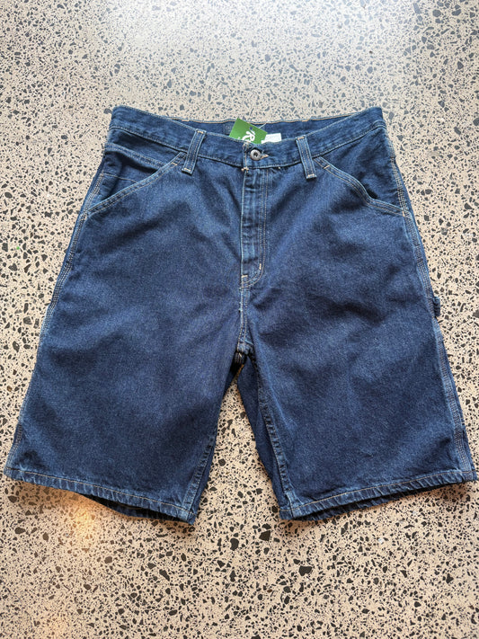 Dark blue wash denim jorts, size 34W, laid on polished concrete background. Knee cut with classic stitching and soft wash