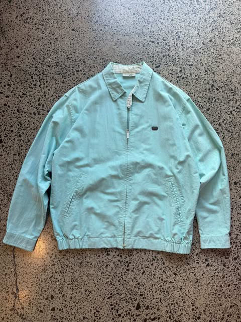 Turquoise Lacoste collared zip-up jacket laid flat on polished concrete background, vintage designer jacket with embroidered crocodile logo and clean fit.