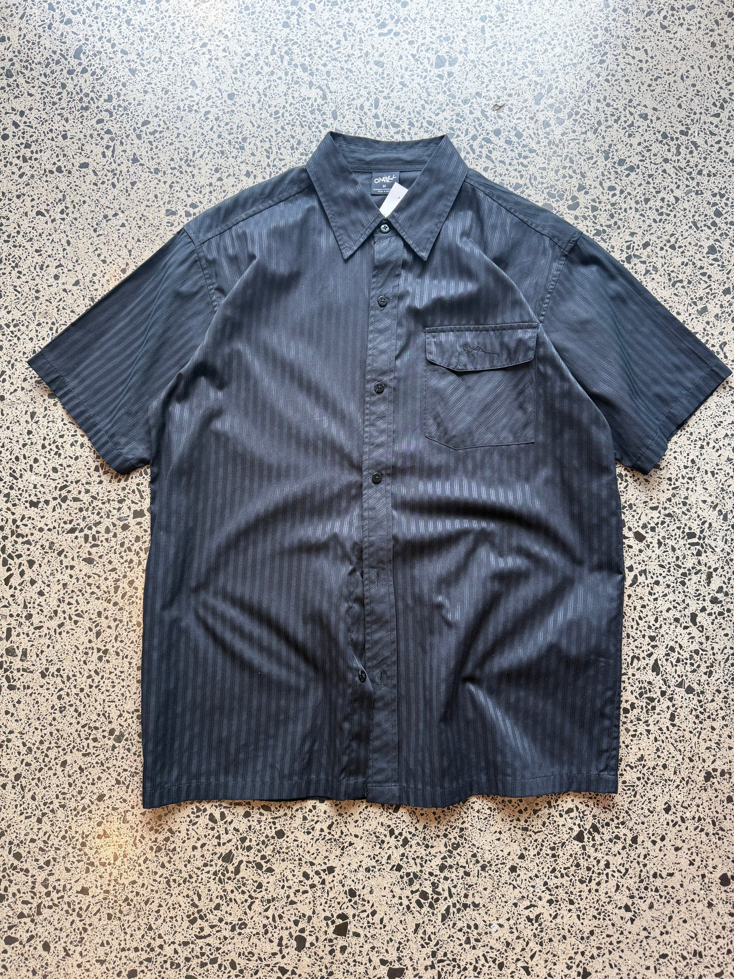O’Neill short-sleeve button-up shirt in size M laid flat on a polished concrete background featuring relaxed surf-style tailoring
