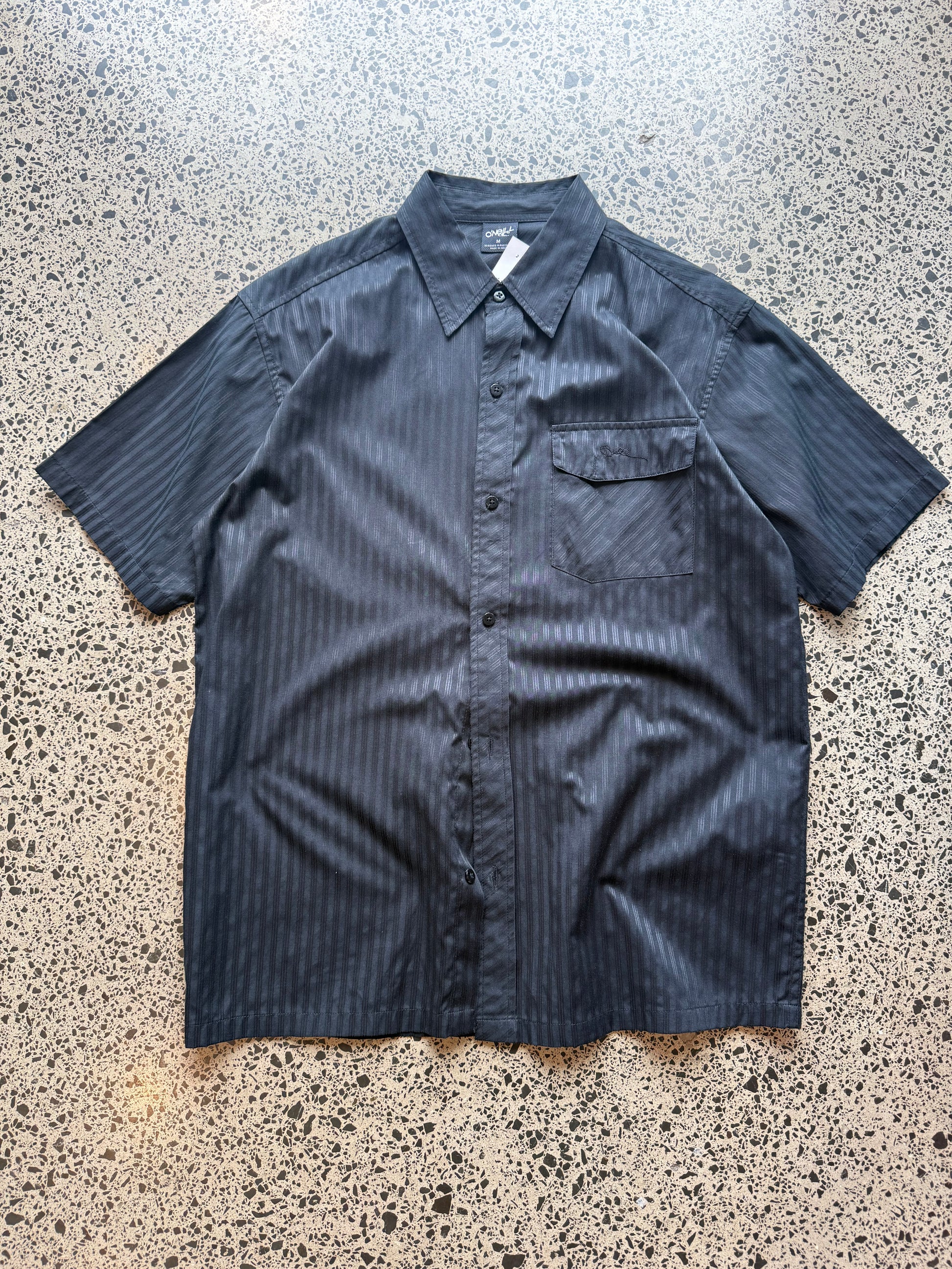 O’Neill short-sleeve button-up shirt in size M laid flat on a polished concrete background featuring relaxed surf-style tailoring