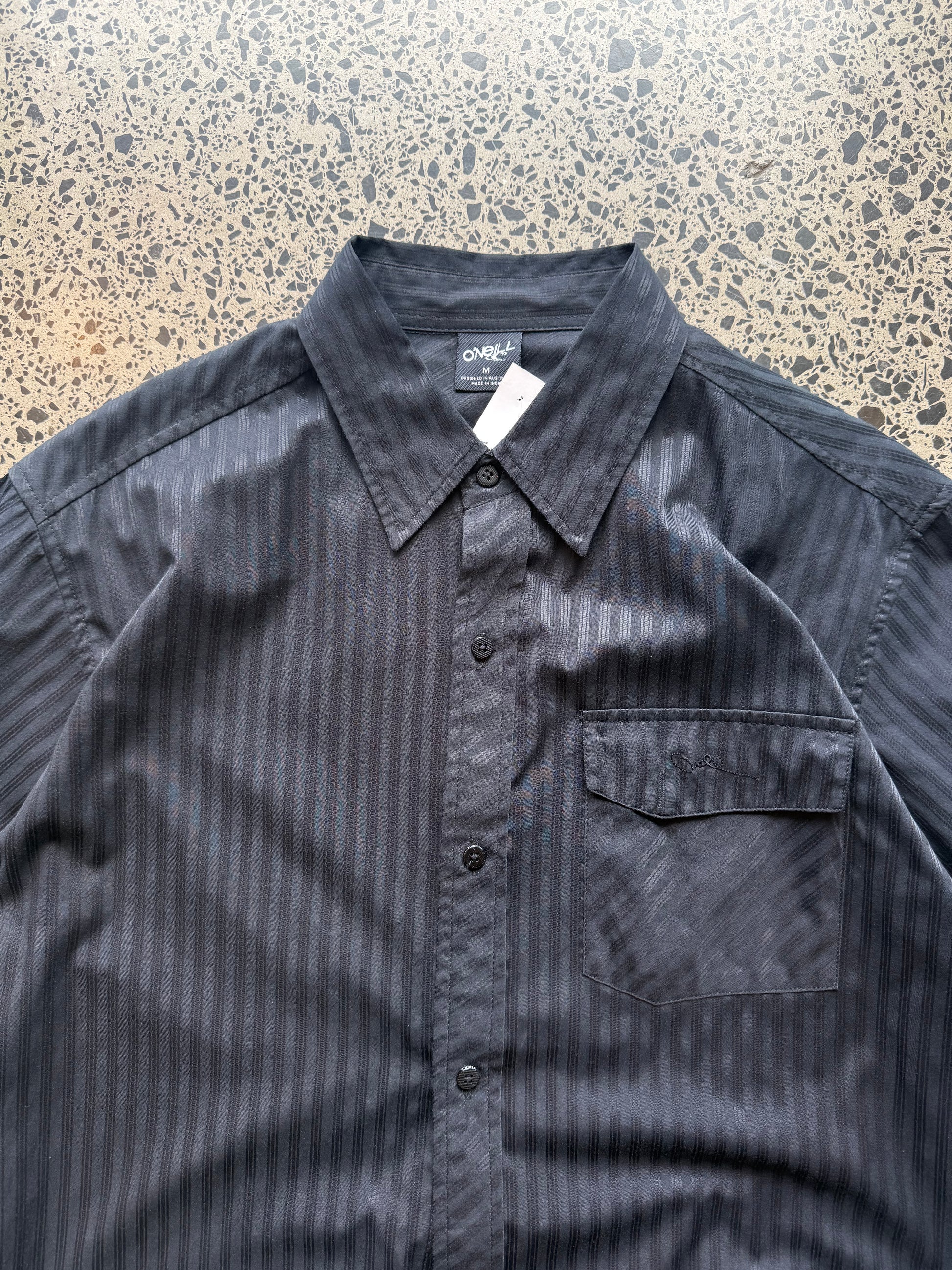 O’Neill short-sleeve button-up shirt in size M laid flat on a polished concrete background featuring relaxed surf-style tailoring