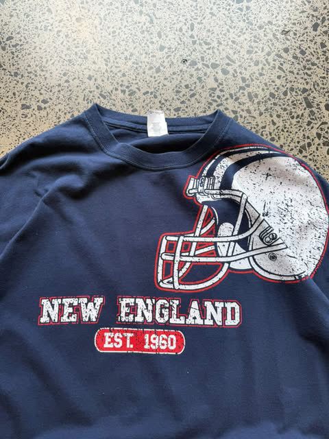Modern New England Patriots NFL T-shirt in size XL laid on a polished concrete background featuring bold team graphics.