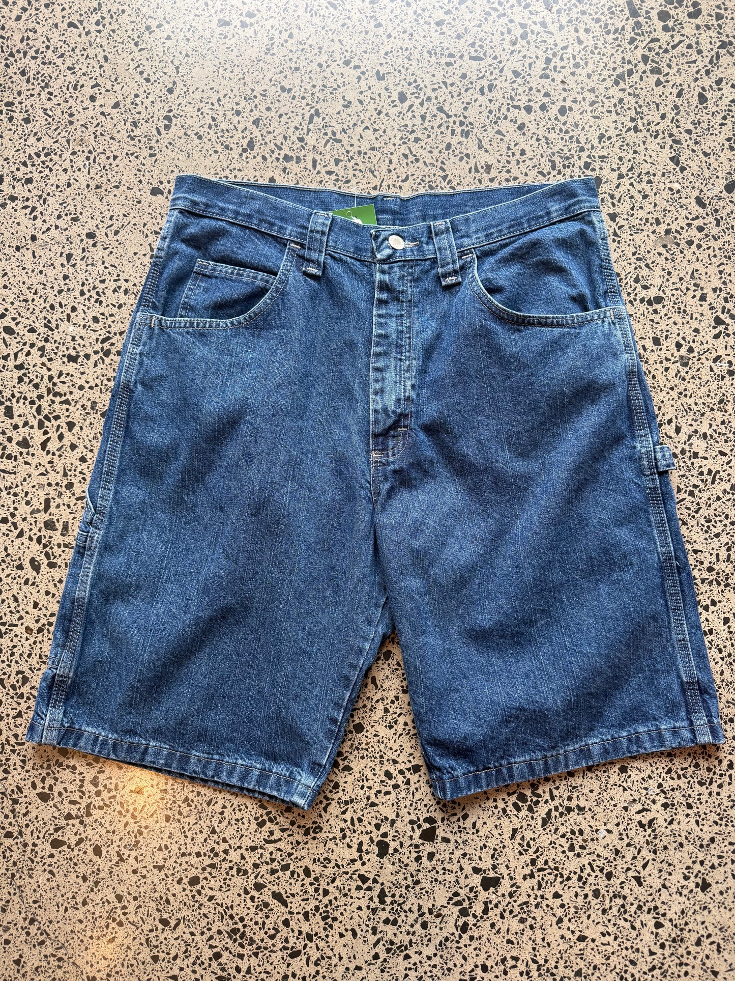 Wrangler Original dark blue wash denim jorts, size 32W, laid on polished concrete background. Knee cut with classic stitching and soft wash
