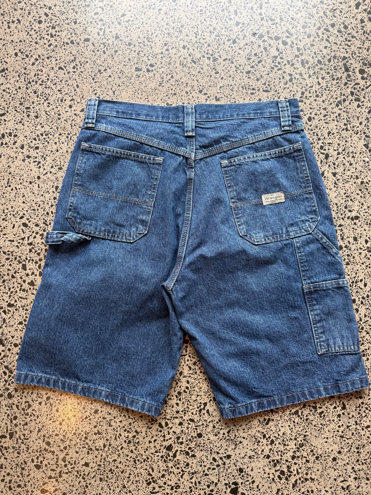 Wrangler Original dark blue wash denim jorts, size 32W, laid on polished concrete background. Knee cut with classic stitching and soft wash
