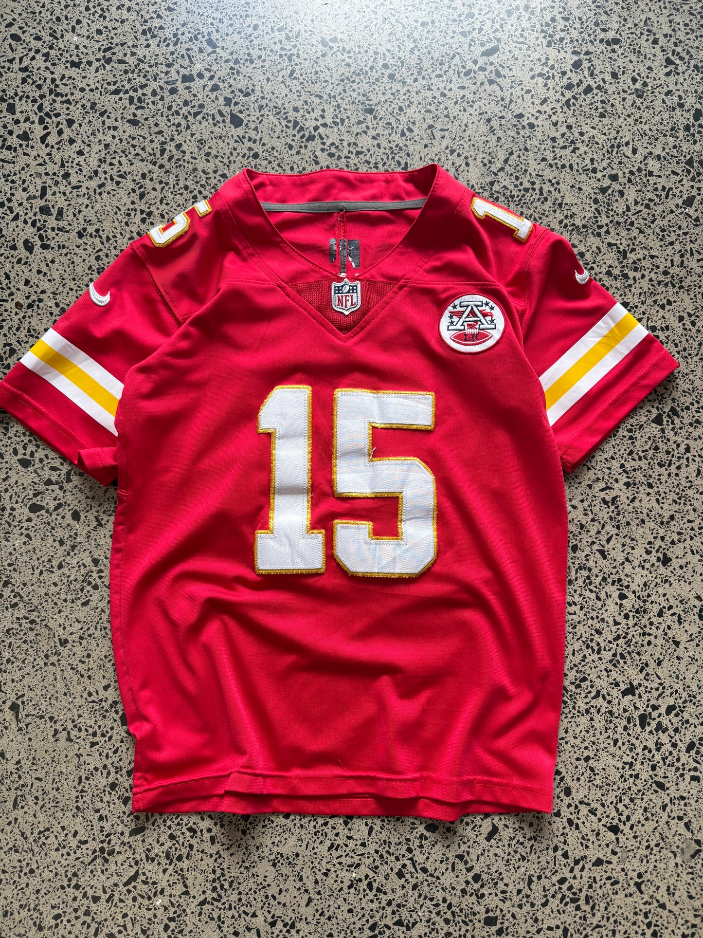 Kansas City Chiefs Patrick Mahomes NFL jersey in Youth Large laid on polished concrete background — red with white and yellow detailing and Chiefs logo.