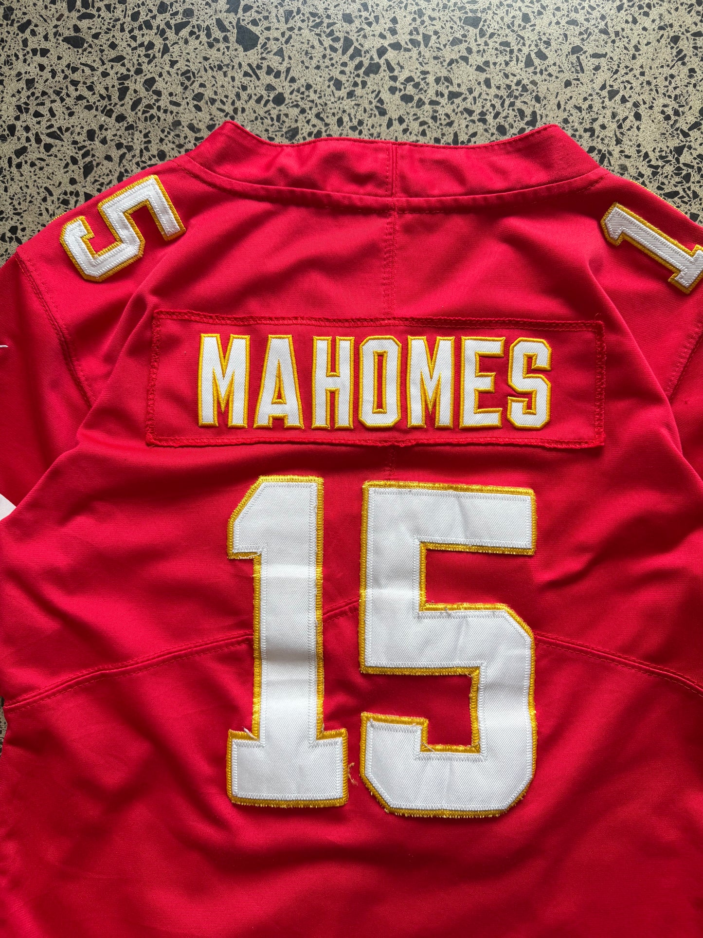 Kansas City Chiefs Patrick Mahomes NFL jersey in Youth Large laid on polished concrete background — red with white and yellow detailing and Chiefs logo.