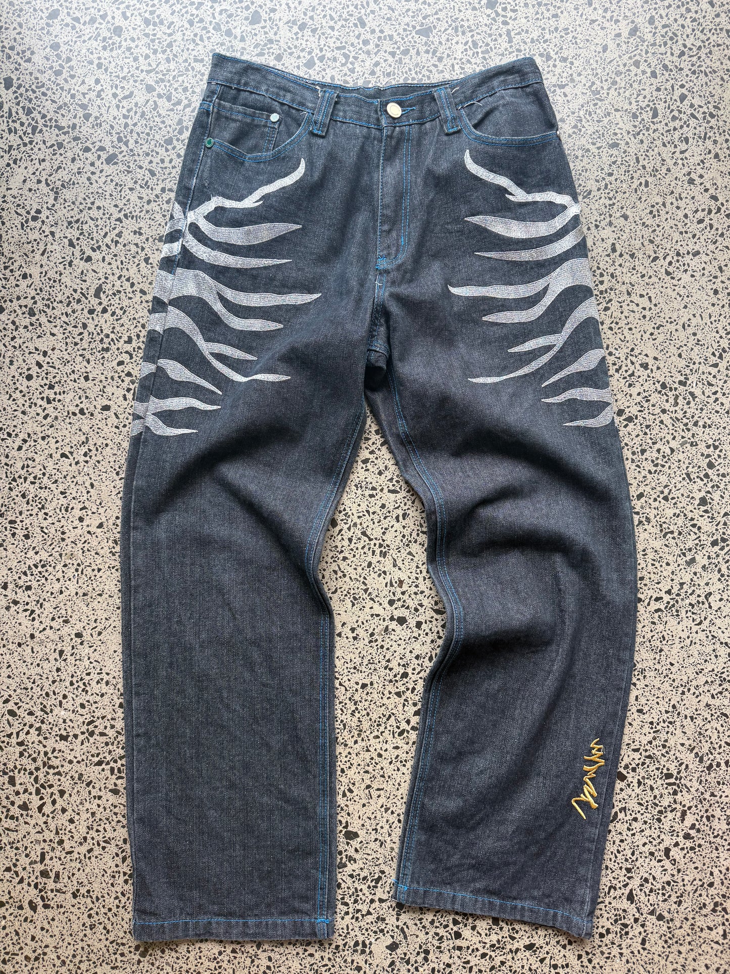 Coogi tiger embroidered denim jeans, 32W x 34L, laid on a polished concrete background — medium-blue denim with bold tiger embroidery and contrast stitching.