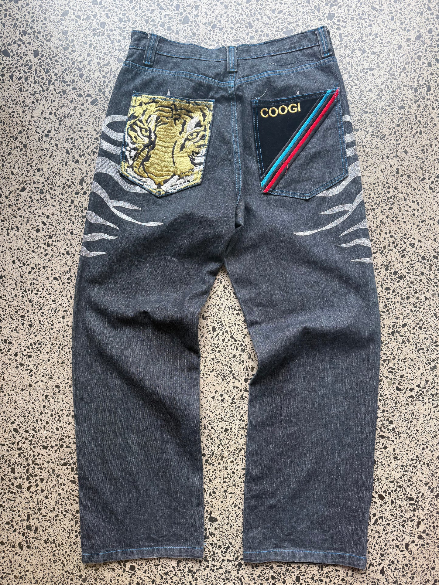Coogi tiger embroidered denim jeans, 32W x 34L, laid on a polished concrete background — medium-blue denim with bold tiger embroidery and contrast stitching.